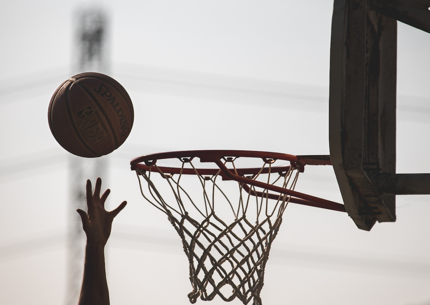 A hand reaches up for a basketball. The basketball is in the air next to a basketball hoop.